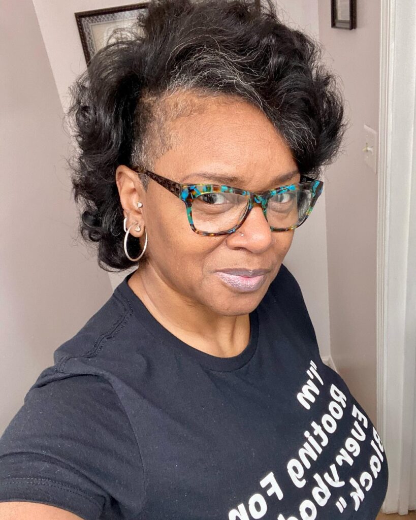 A woman wearing a bold asymmetrical hairstyle with one side shaved and the rest of her dark curly hair styled with volume.
