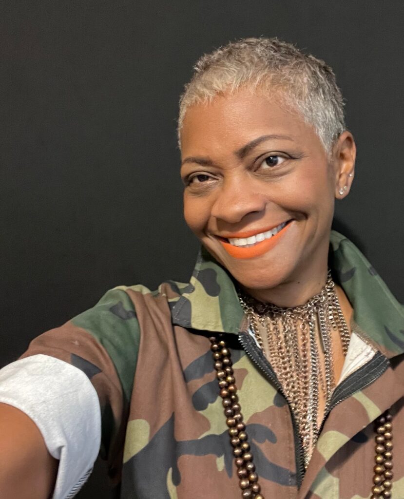 A smiling Black woman over 60 with an ultra-short silver buzz cut wearing a camouflage jacket and layered necklaces.