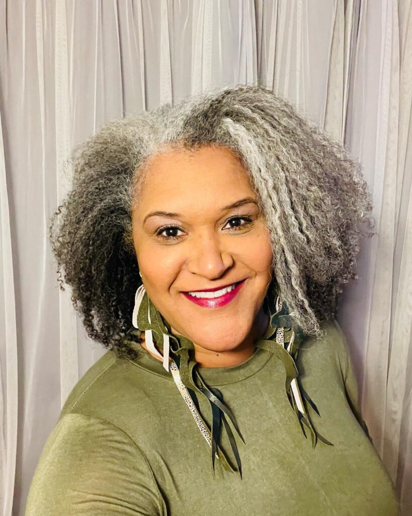A smiling woman over 60 with a voluminous salt-and-pepper curly bob and natural textured silver hair.
