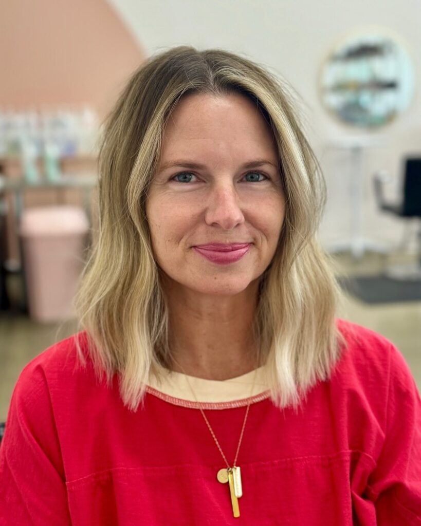 A woman with a round face wearing a medium-length textured blonde lob with dark roots and bright face-highlighting pieces.