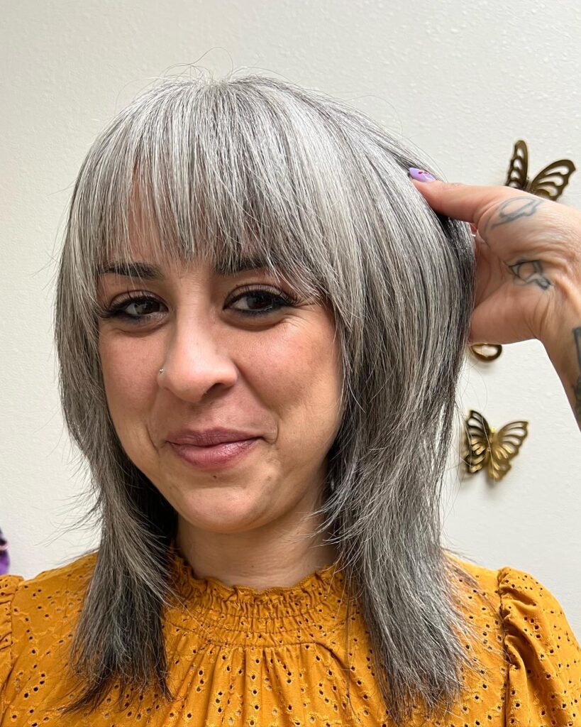 A close-up of a woman showing off a shoulder-length silver shag haircut with full straight bangs and textured layers.