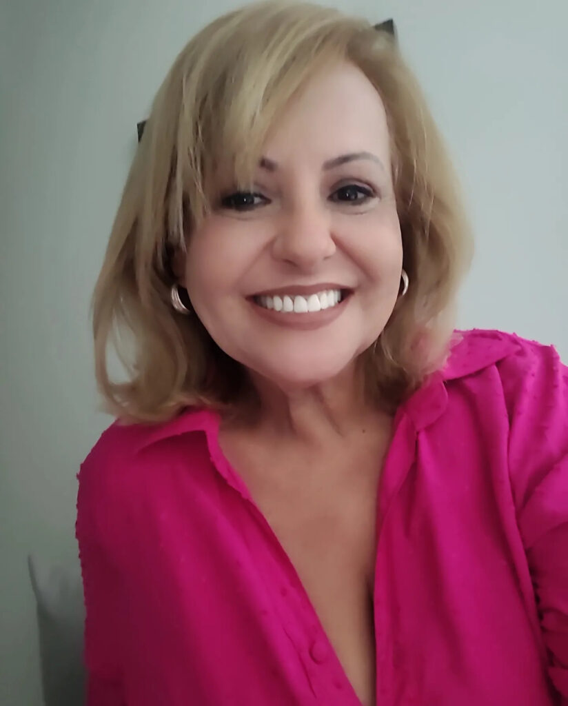 A woman with a round face smiling brightly and wearing a warm, light blonde shoulder-length bob with layers and a swept fringe.