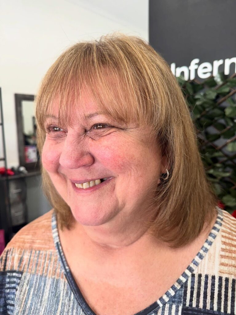 A smiling woman with a round face wearing a medium-length golden bronde hairstyle with full wispy bangs and soft layers.
