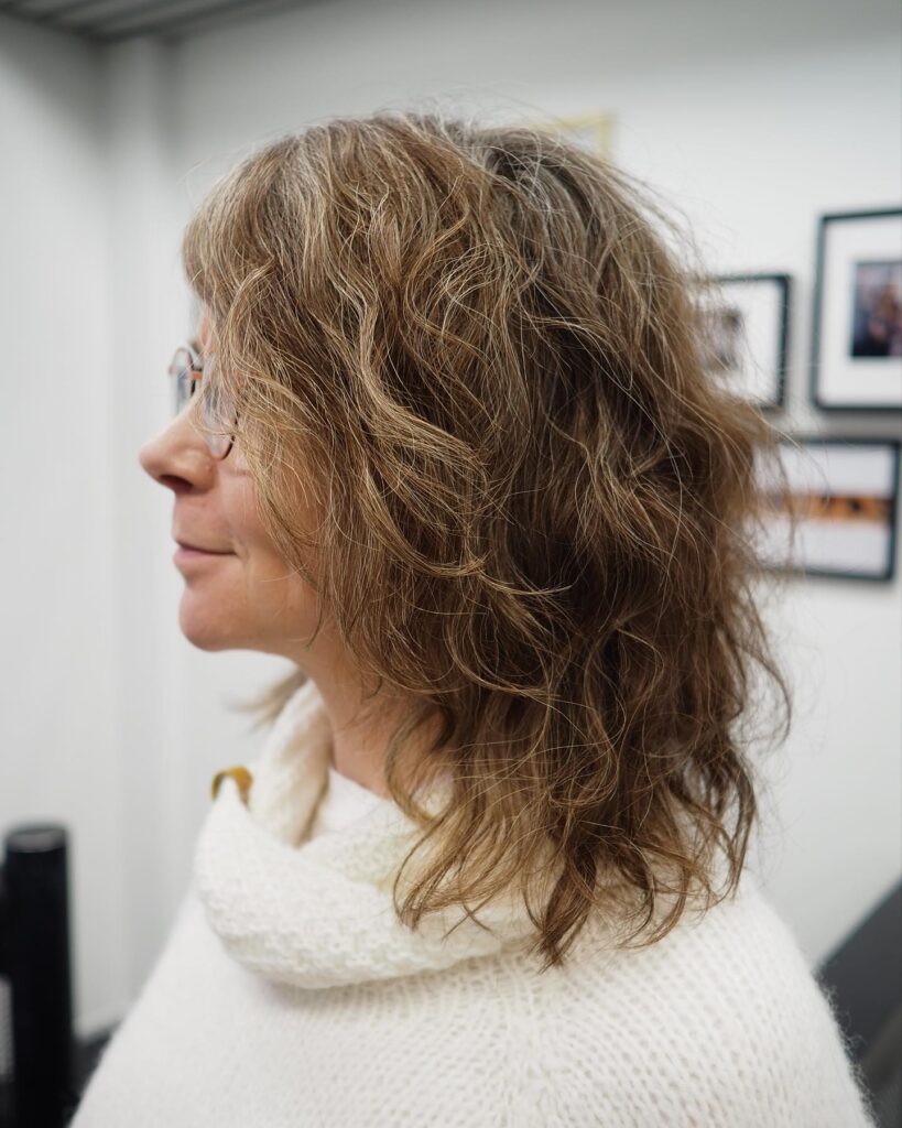 A woman in profile with a round face wearing a medium-length shaggy hairstyle with messy waves and dimensional blonde and grey tones.