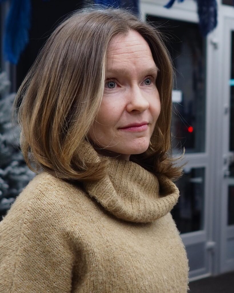 A woman with a round face wearing a medium-length rounded brunette lob with warm caramel highlights and inward-curved ends.