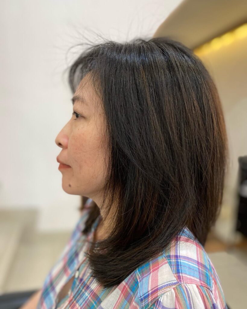 A woman in profile with a round face wearing a sleek, medium-length espresso brown layered lob with ends that curve inward.