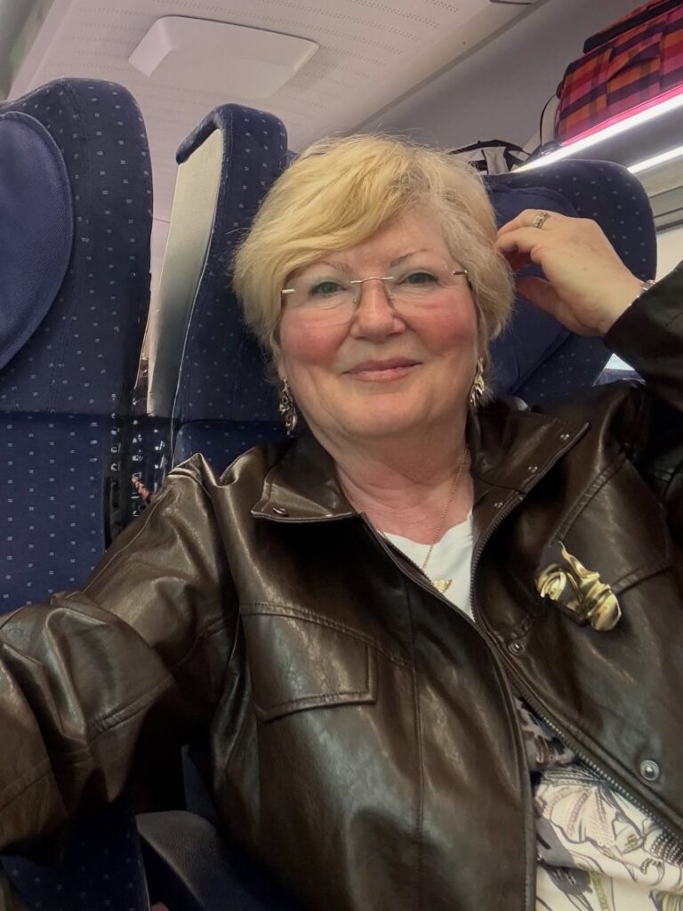 A woman over 60 with a layered golden blonde pixie hairstyle and side-swept bangs wearing a brown leather jacket.