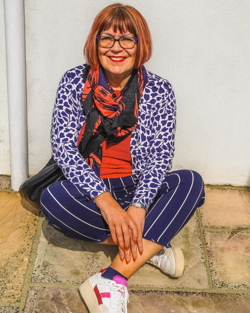 A woman over 60 with a vibrant copper bob and blunt bangs sitting cross-legged outdoors.