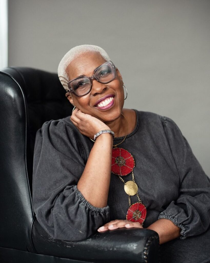 A woman over 60 with a stylish platinum blonde buzz cut featuring an etched side design and wearing large glasses.