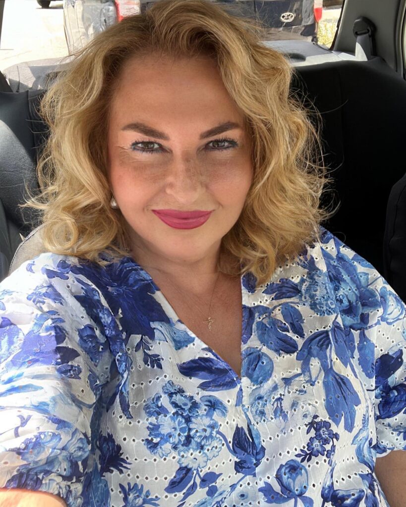 A woman over 60 with shoulder-length honey-blonde wavy hair wearing a blue floral top.
