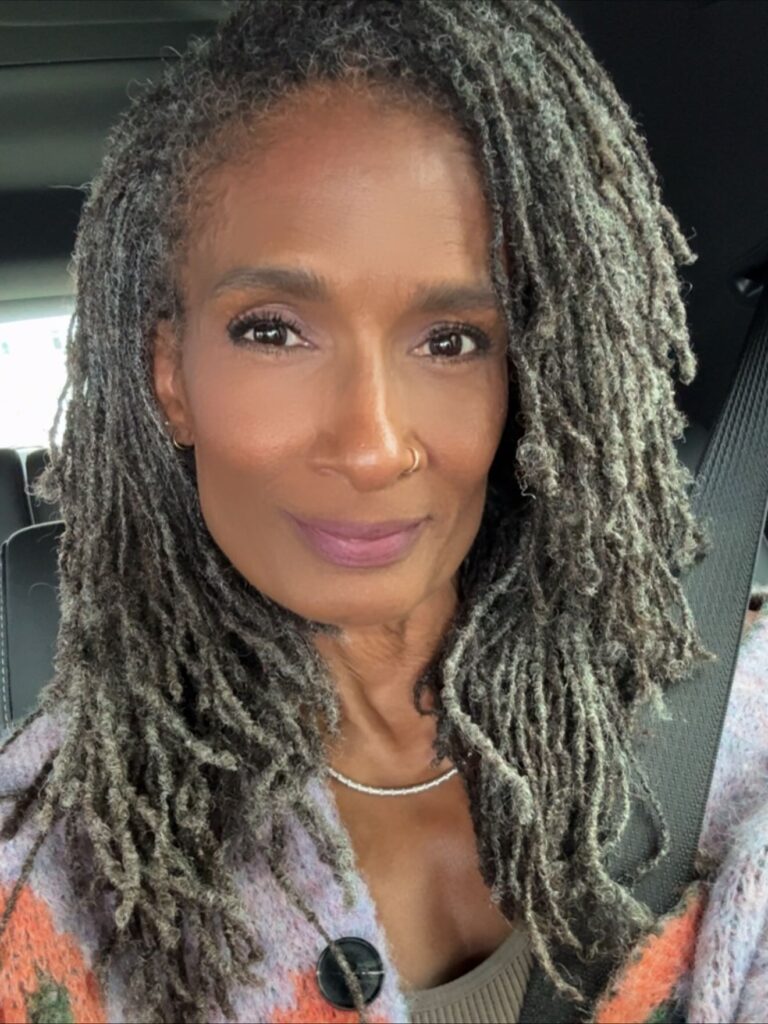 A woman over 60 with long, slender salt-and-pepper sisterlocks styled with a soft side part.