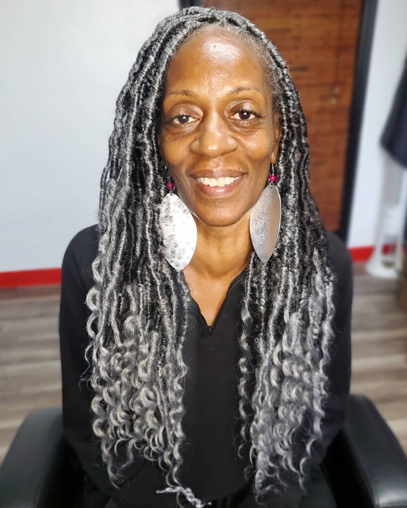A woman over 60 with long salt-and-pepper goddess locs featuring wavy ends and a middle part.