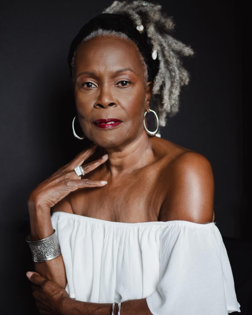 A woman over 60 with long, silver natural locs gathered to the side and styled with a black velvet headwrap and shell accessories.