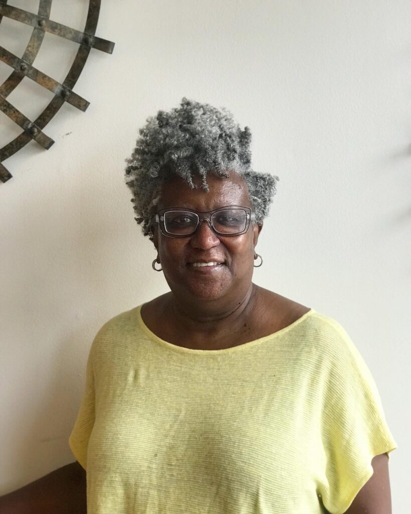 A smiling woman with a short, tapered salt-and-pepper frohawk featuring tight natural curls with height at the center.