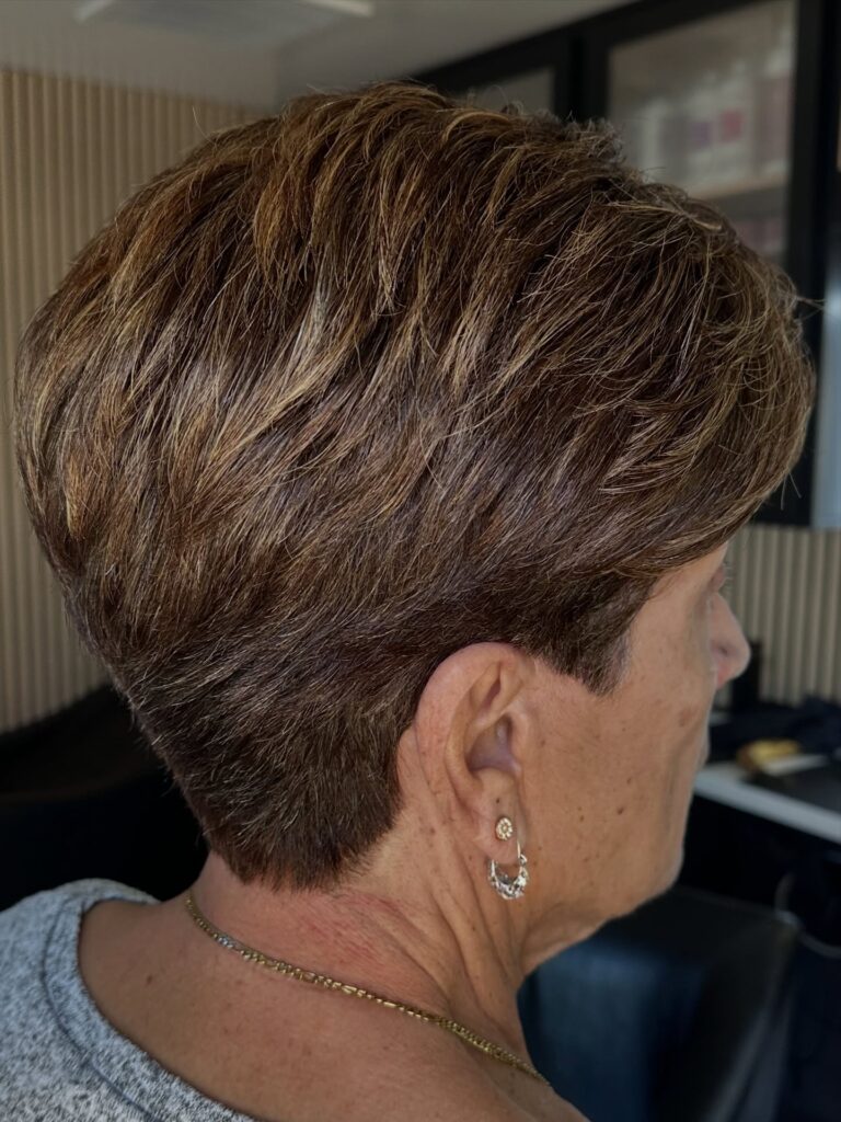 A woman over 60 is seen from the side with a short, tapered pixie cut in dark brown with bronze highlights.