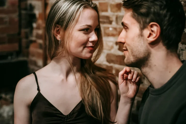 A couple shares an intimate moment, smiling at each other against a rustic brick wall. The woman has long, wavy blonde hair, while the man has short, dark hair.