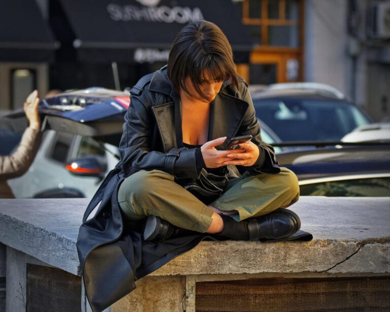 The Kind of Text Chemistry You Can Feel Right Away A woman with a sleek bob hairstyle sits cross-legged on a concrete ledge, wearing a black leather coat and olive green pants, focused on her phone.