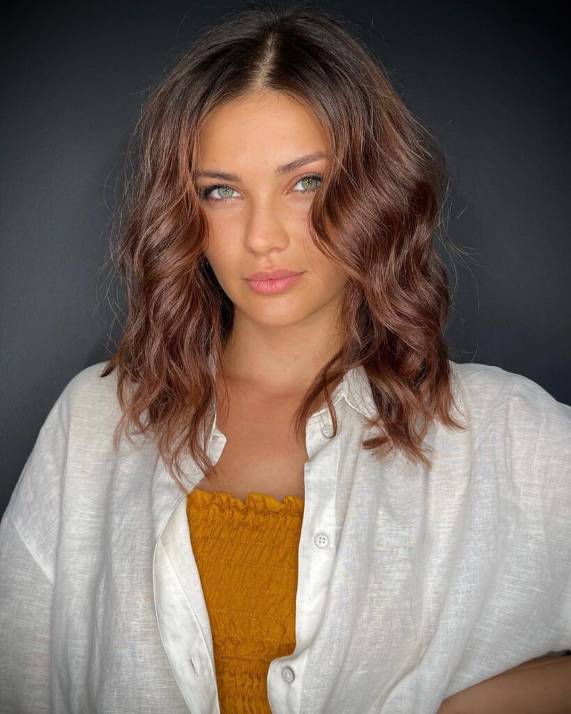A woman with green eyes looking forward wearing a warm chestnut brown shoulder-length choppy bob hairstyle with loose waves and a center part.