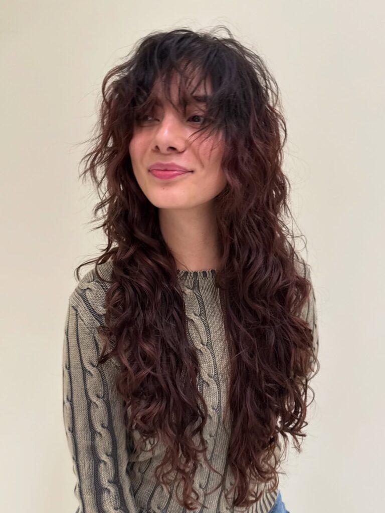 A young woman looking to the side with a smile, wearing a dark mahogany brown long choppy lob hairstyle with voluminous shaggy layers and wispy bangs.