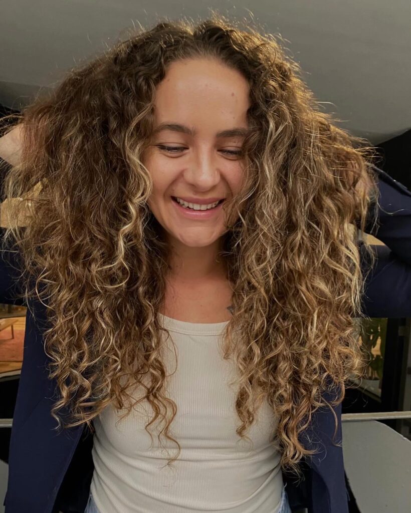 A smiling woman showing off a long, highly voluminous curly choppy bob hairstyle with honey blonde highlights and face-framing layers.