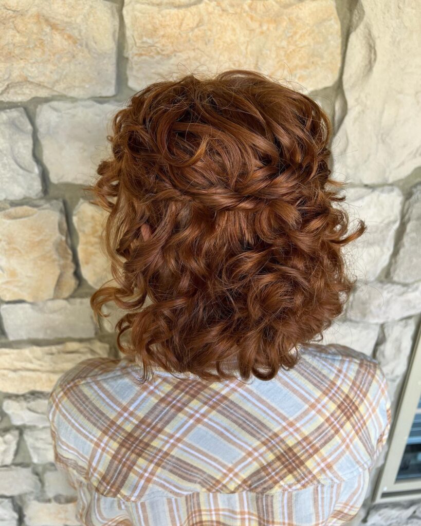 A back view of a woman with an auburn red neck-length choppy bob hairstyle styled into a half-up look with bouncy natural curls.