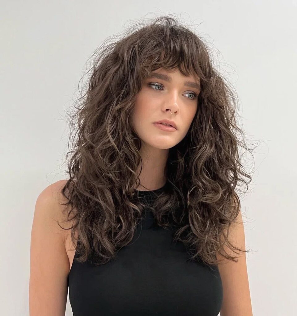 A woman with a textured ash brown shoulder-length choppy bob hairstyle featuring voluminous wavy layers and a full face-framing fringe.