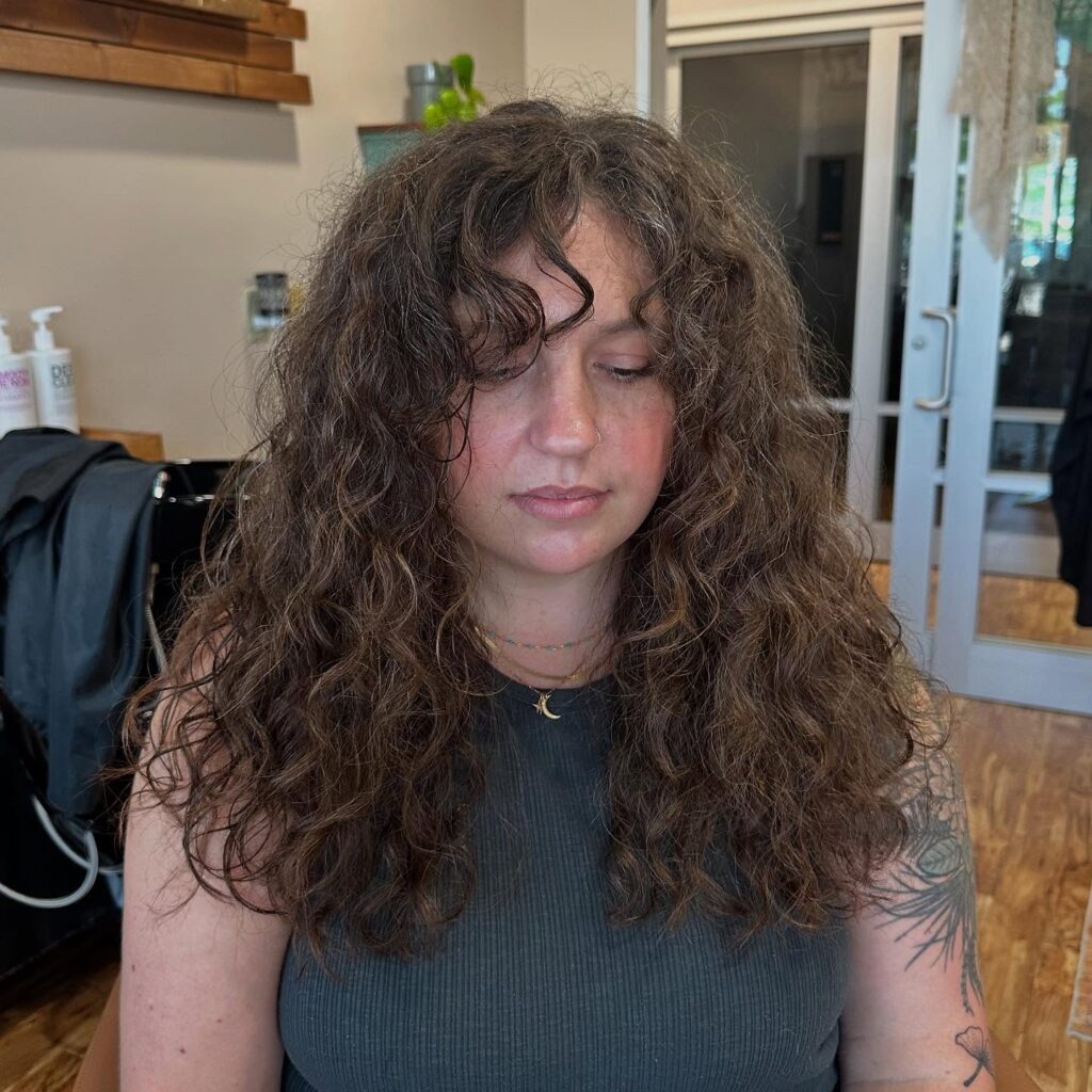 A woman with tattoos looking down, showing off a voluminous brown shoulder-length choppy lob hairstyle with natural curls and wispy bangs.
