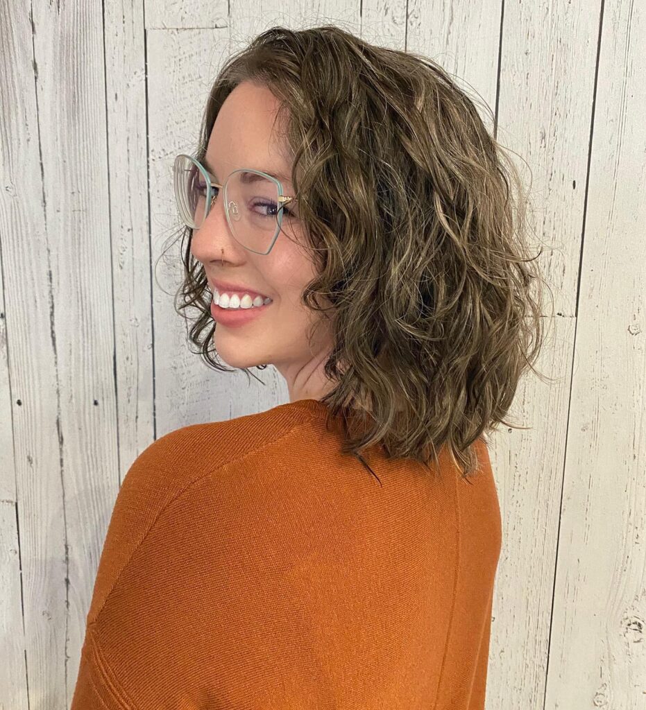 A smiling woman wearing glasses looks over her shoulder to show off a sandy bronde neck-length choppy bob with relaxed tousled waves.