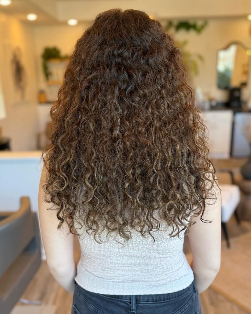A back view of a woman with long highlighted brown and golden blonde curly hair cut into a tapered V-shaped choppy lob.