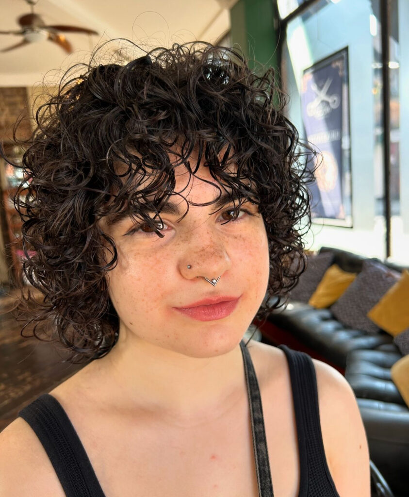 A young woman with freckles and nose piercings wearing a glossy black chin-length choppy curly bob hairstyle with wet-look ringlet bangs.