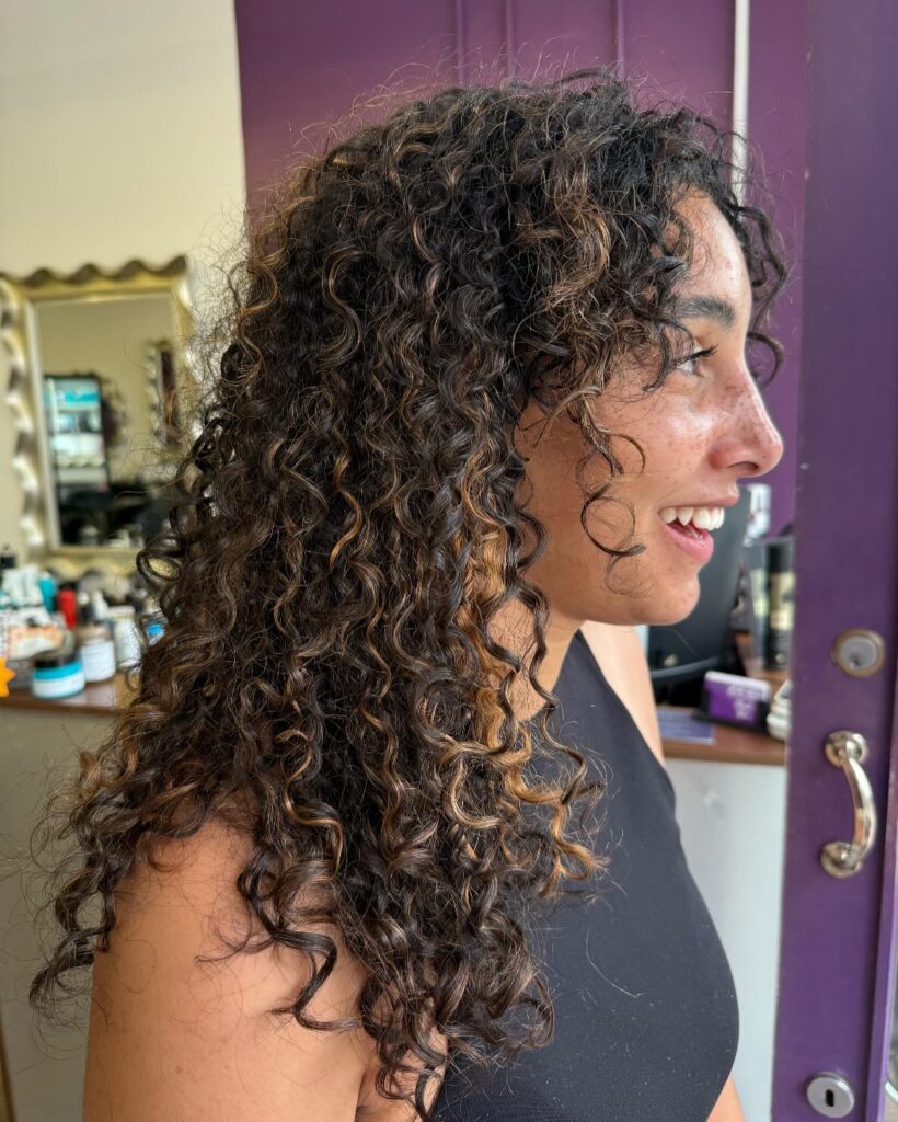 A side profile of a woman smiling with a shoulder length curly choppy bob hairstyle featuring dark brown ringlets and warm caramel highlights.