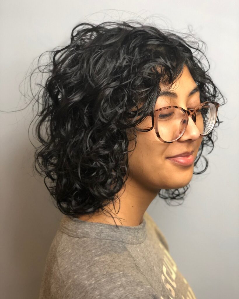 A side profile view of a smiling woman wearing glasses and a jet black neck-length choppy curly bob hairstyle with glossy defined ringlets.