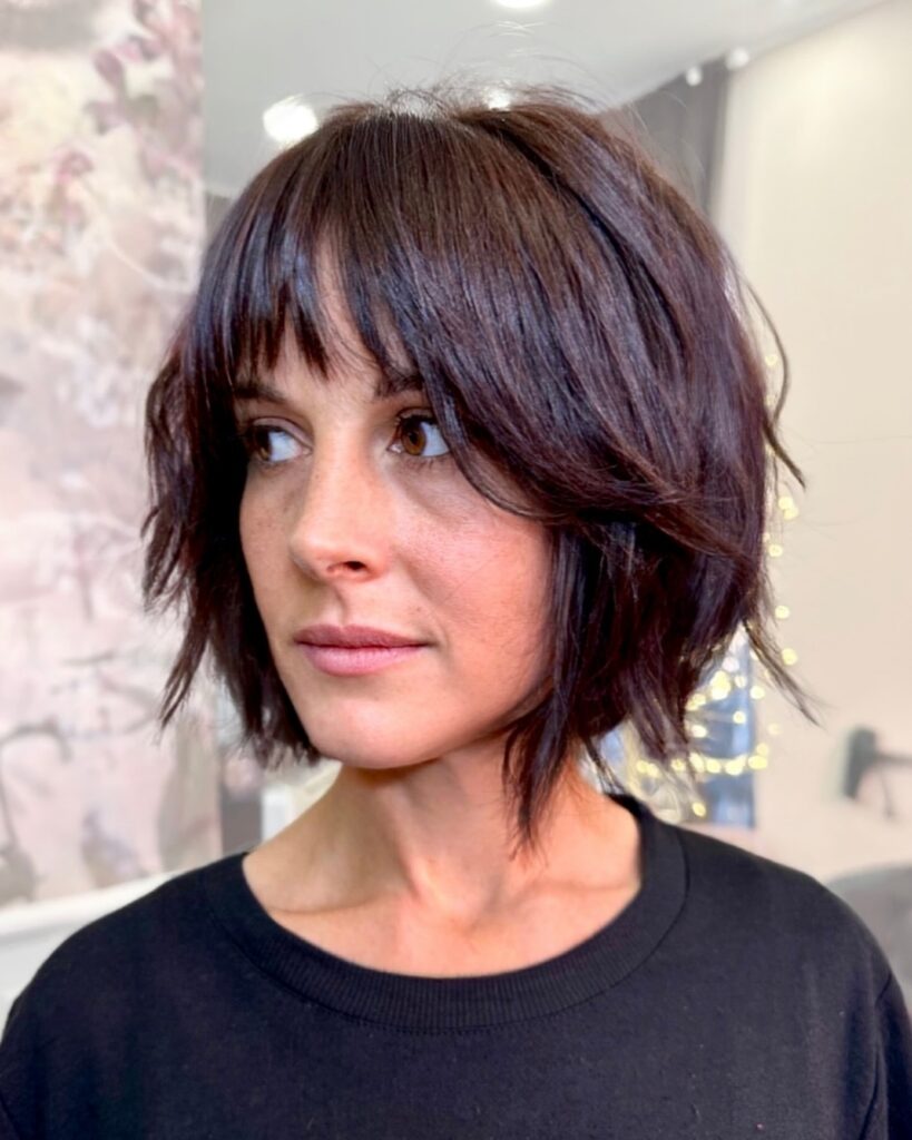 A woman with a chin-length shaggy dark brunette choppy bob featuring wispy straight bangs and textured piecey layers.