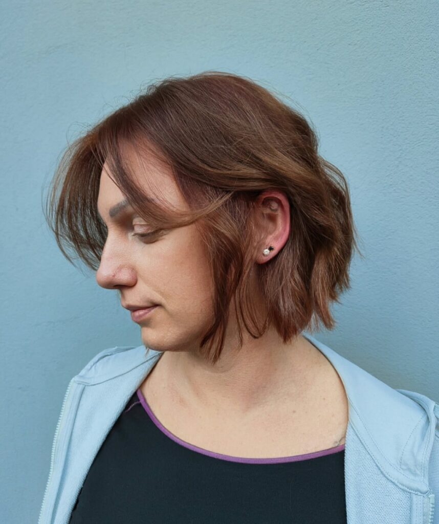 A woman with a chin-length auburn choppy bob showing long wispy curtain bangs tucked behind the ear.