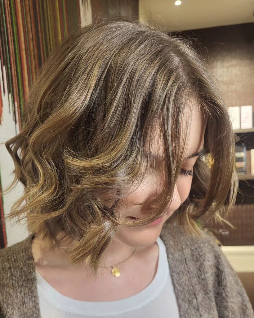 A woman looking down to show a chin-length choppy honey bronde bob with bouncy waves and long face-framing curtain bangs.