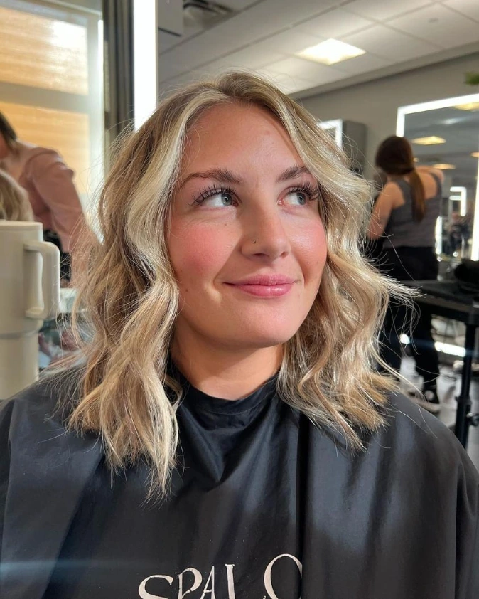A woman with a shoulder-length textured caramel blonde bob featuring relaxed waves and thick face-framing curtain bangs.