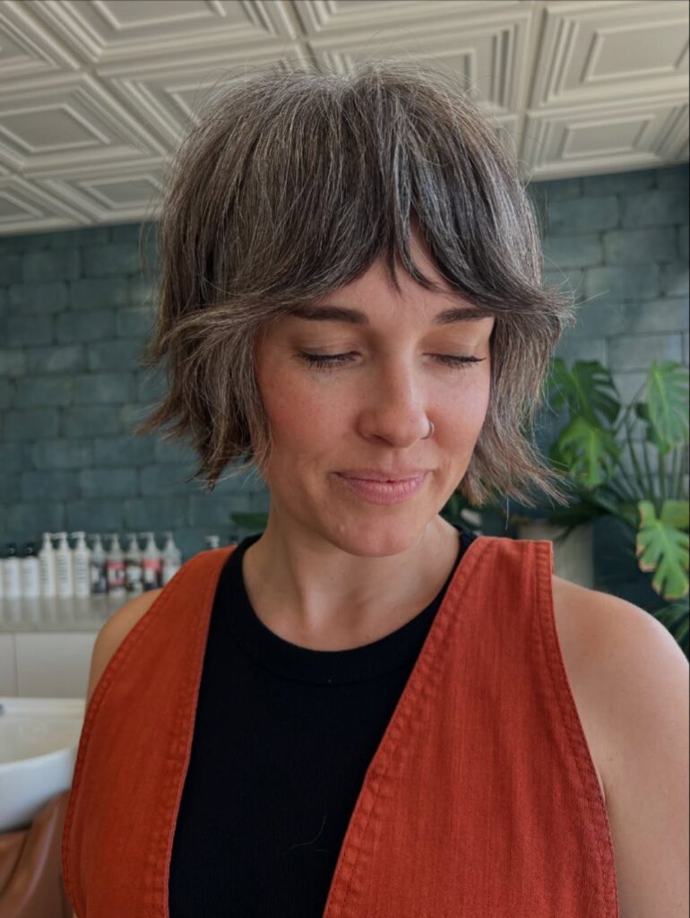 A woman with a jaw-length salt and pepper choppy bob featuring middle-parted curtain bangs and shaggy flipped-out layers.