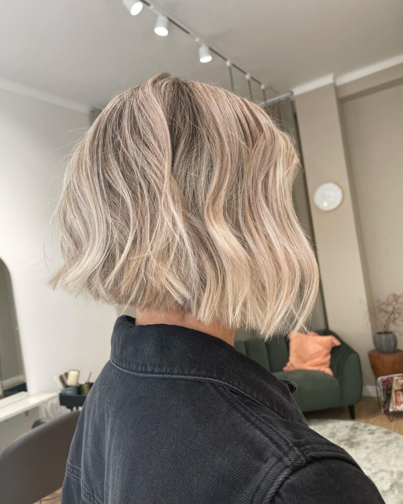 A woman viewed from behind and the side showing a chin-length choppy ash blonde bob with blunt edges and soft texture.