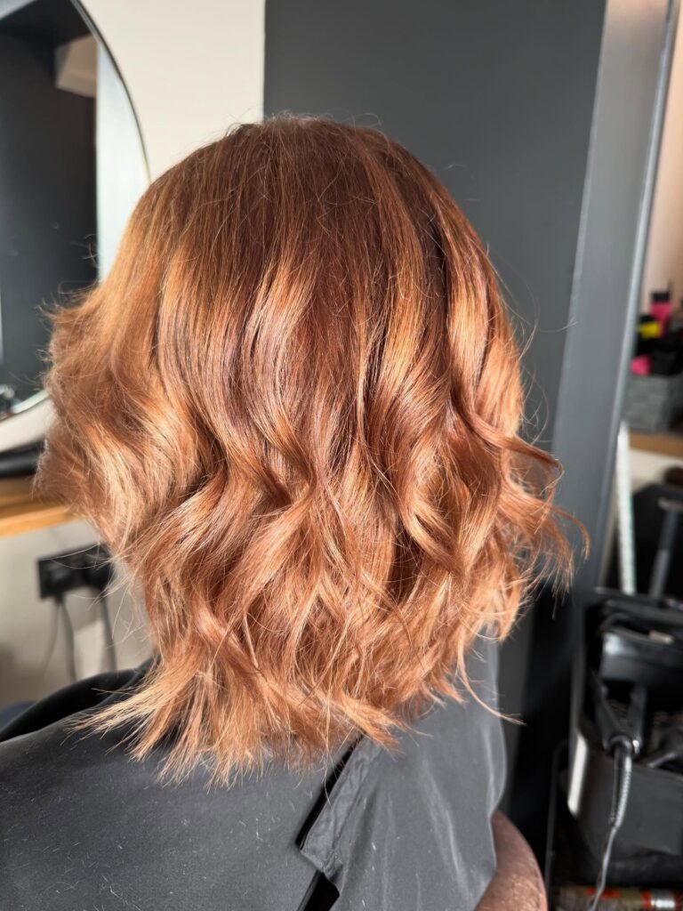 A back view of a woman with a shoulder-length choppy copper lob featuring textured beachy waves and wispy layers.
