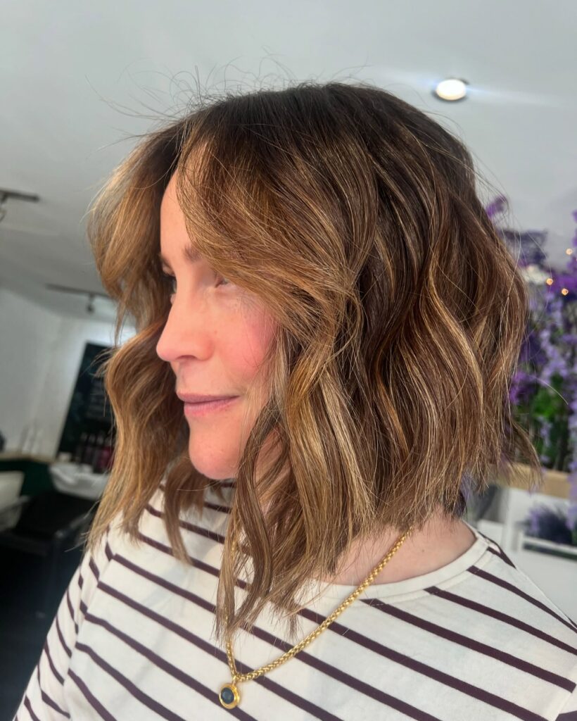 A woman with a collarbone-length choppy brunette lob featuring warm caramel highlights, textured waves, and voluminous curtain bangs.