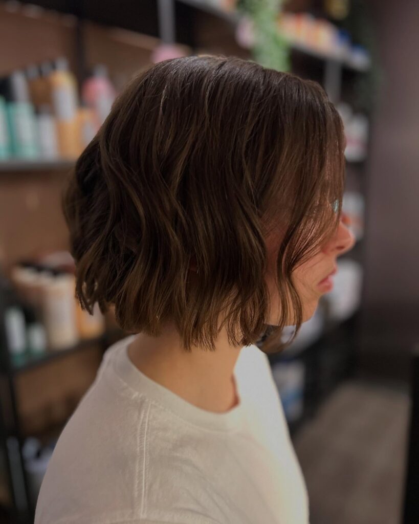 A side profile view of a woman with a chin-length choppy brunette bob featuring soft lived-in waves and textured face-framing layers.