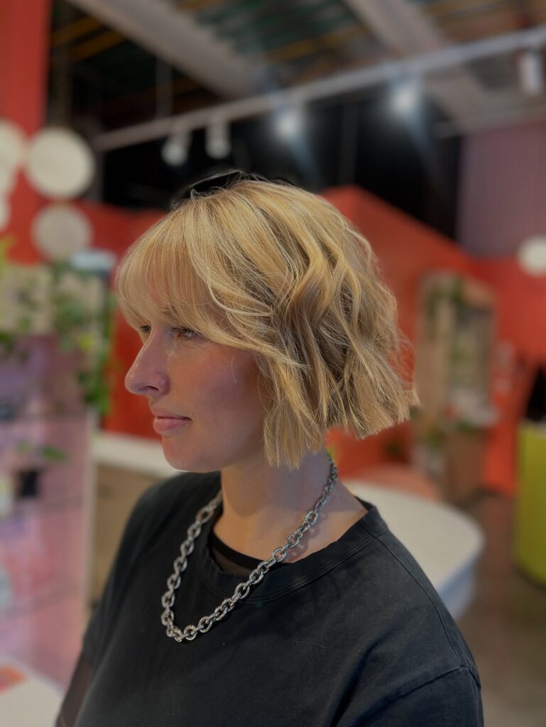 A woman with a jaw-length choppy blonde bob featuring soft curtain bangs and textured beachy waves.