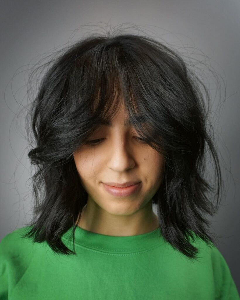 A woman looking down showcasing a messy jet black shag bob with voluminous choppy layers and curtain bangs.