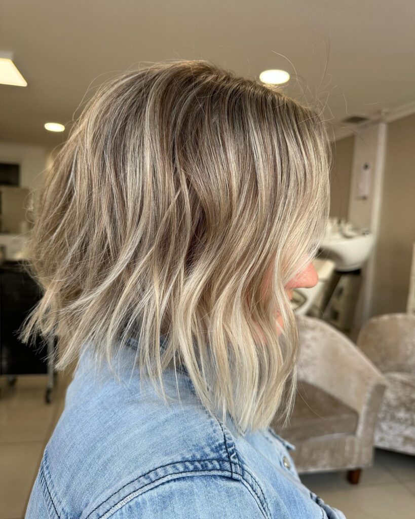 A side profile of a woman wearing a textured blonde lob with dark roots and choppy ends styled in loose waves