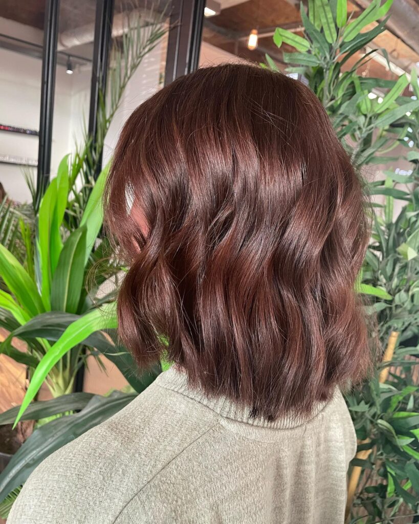 The back and side view of a woman showcasing a glossy chestnut brown shoulder-length bob styled with soft, loose waves.