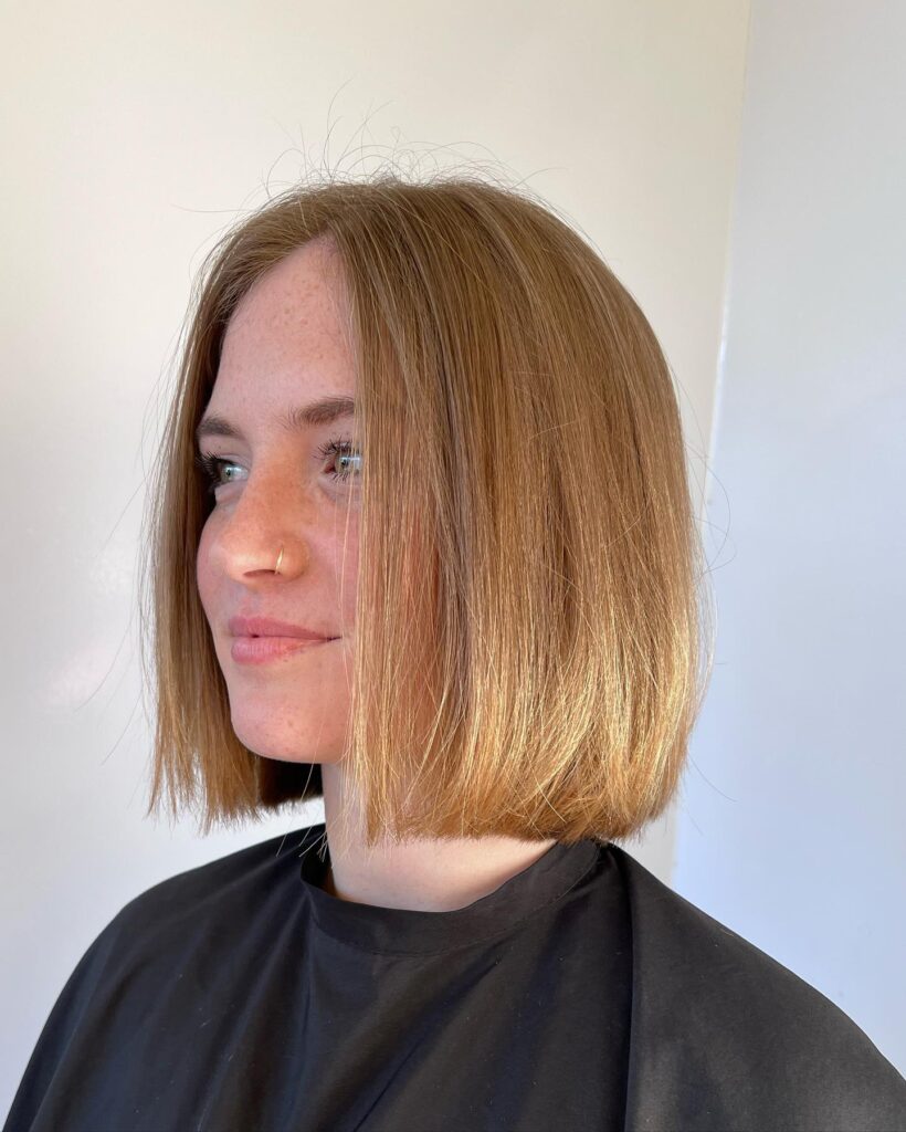 A side profile of a woman showcasing a sleek chin length golden honey blonde bob with texturized ends and a middle part.