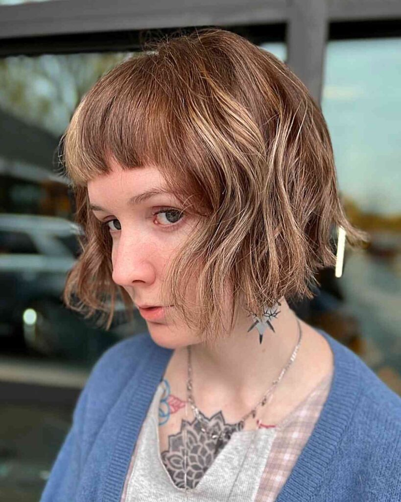 A side profile view of a woman with a chin length caramel French bob featuring piecey texture and blunt micro bangs.