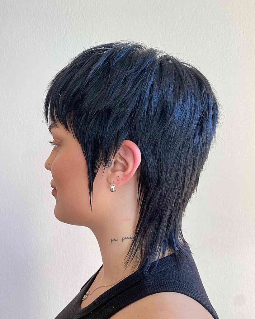 A side profile of a woman with a short, heavily layered black shaggy bob featuring subtle blue highlights and a longer wispy back.