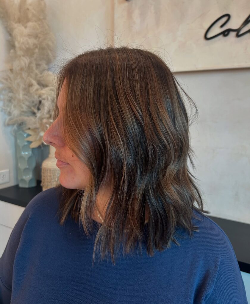 A side profile view of a woman with a collarbone length dark brunette lob featuring soft caramel highlights and loose textured waves.