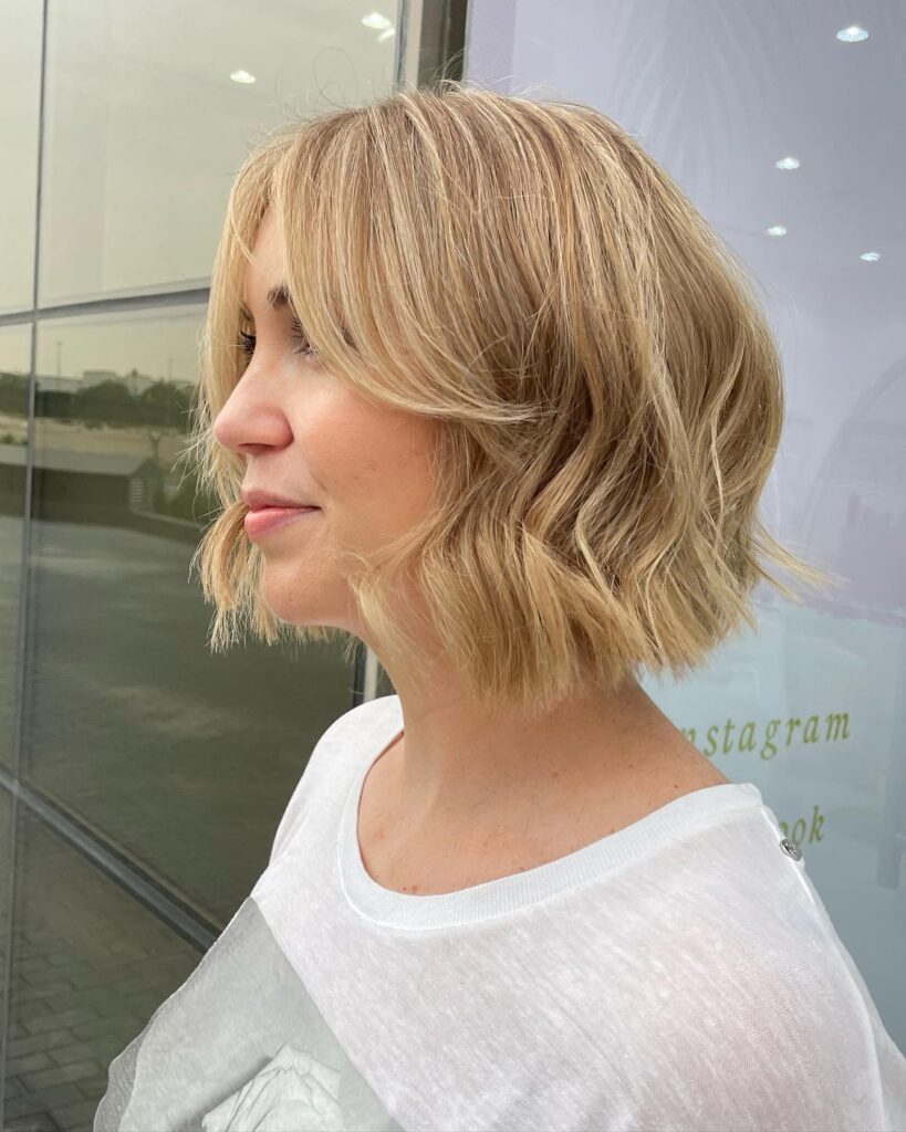 A side profile of a woman showcasing a neck length honey blonde bob with soft waves and wispy curtain bangs.